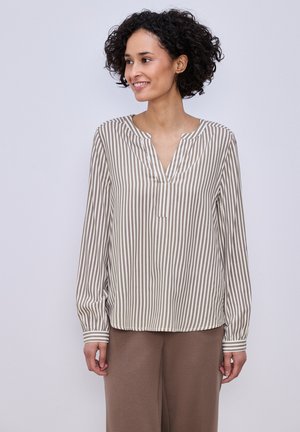 Frau mit lockigem Haar, die eine beige-weiße, vertikal gestreifte Langarmbluse und braune Hose trägt, lächelt und schaut zur Seite.