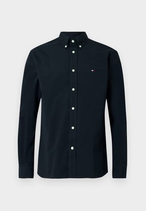 Chemise à manches longues bleu marine foncé avec col boutonné, dotée de boutons blancs et d'un petit logo tricolore sur la poitrine gauche. Tissu en coton.