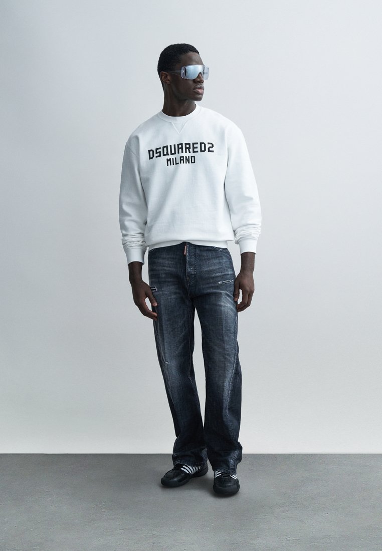 Sweat-shirt blanc avec "DSQUARED2 MILANO" en noir, associé à un jean denim foncé et des chaussures noires avec des bandes blanches. Le modèle porte des lunettes.