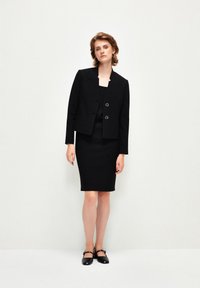 Czarna dopasowana marynarka z dwoma okrągłymi guzikami, zestawiona z dopasowaną czarną spódnicą i pasującymi czarnymi butami. Czyste linie i strukturalny design.