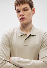 Bež dolg rokav polo majica z teksturiranim materialom, z ovratnikom in majhno etiketo z logotipom "BOSS" na prsih.