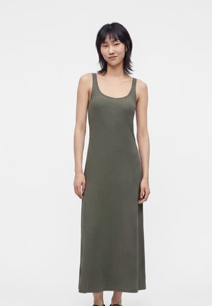 Femme debout et souriante, portant une robe débardeur sans manches, vert olive, longue jusqu’aux chevilles, sur fond blanc uni.