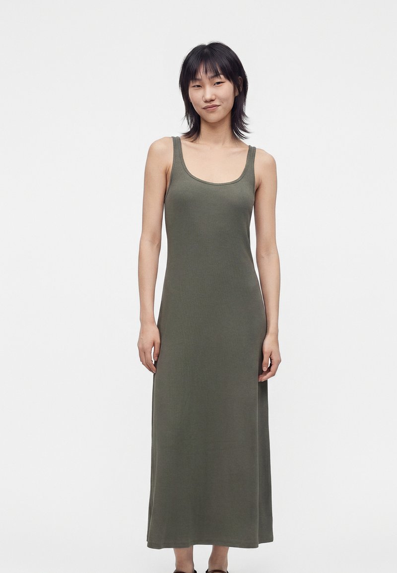 Femme debout et souriante, portant une robe débardeur sans manches, vert olive, longue jusqu’aux chevilles, sur fond blanc uni.