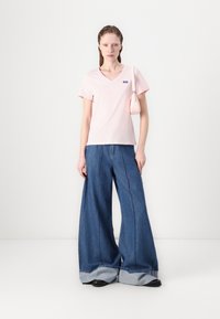 Lys rosa v-hals t-skjorte, kombinert med brede, blå denimjeans med synlige sømmer. Modellen holder en liten rosa håndveske.