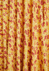 Puuvillakangas keltaisella taustalla, jossa on punavalkoinen kukkakuvio. Kangas on teksturoitu ja laskostettu.