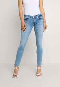 Žena v světle modrých skinny džínách s předními kapsami a bílých sandálech s podpatky, stojící na jednobarevném pozadí.