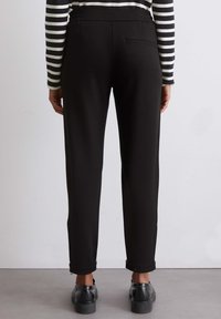 Zwarte tapered broeken met een elastische tailleband, een achterzak en opgerolde manchetten, gecombineerd met zwarte schoenen op een neutrale achtergrond.