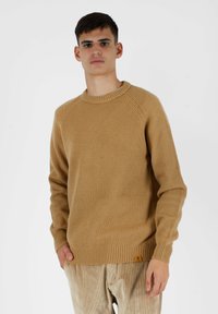 Pullover in maglia a coste di colore beige con scollo rotondo, maniche lunghe a raglan e un piccolo patch con logo all'orlo. Realizzato in un materiale morbido e текстurizzato.