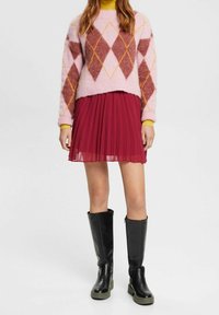 Strikket lyserød sweater med diamantmønstre i burgunder og gul, parret med en plisseret burgunder nederdel og sorte knæhøje støvler med grønne såler.