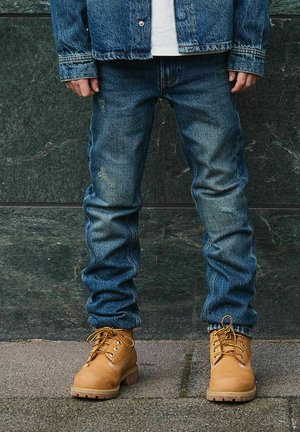 Donkerblauwe denim jeans met lichte vervaging, gecombineerd met tan werkschoenen met gele veters en dikke zolen. Over het geheel een casual outfit.