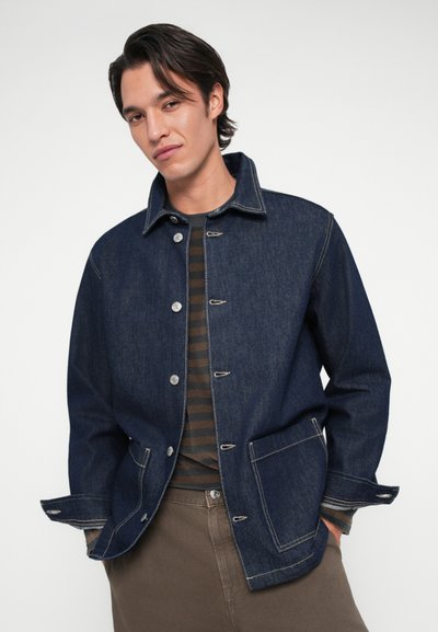 Veste en jean bleu foncé avec un col, des boutons-pression argentés, deux poches avant et des coutures blanches, portée sur une chemise rayée marron.