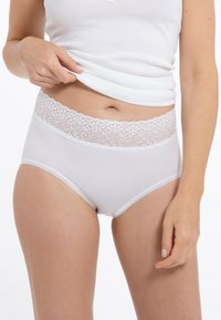 Frau hebt weißes Tanktop an, um weiße Unterwäsche mit Spitzenrand und hohem Taillenschnitt auf heller Haut vor einem weißen Hintergrund zu zeigen.