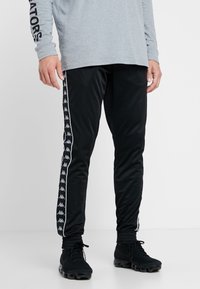Svarta träningbyxor med elastiska muddar, med en vit sidremsa prydd med Kappa-logotyper. Tillverkade av ett mjukt, lätt tyg.