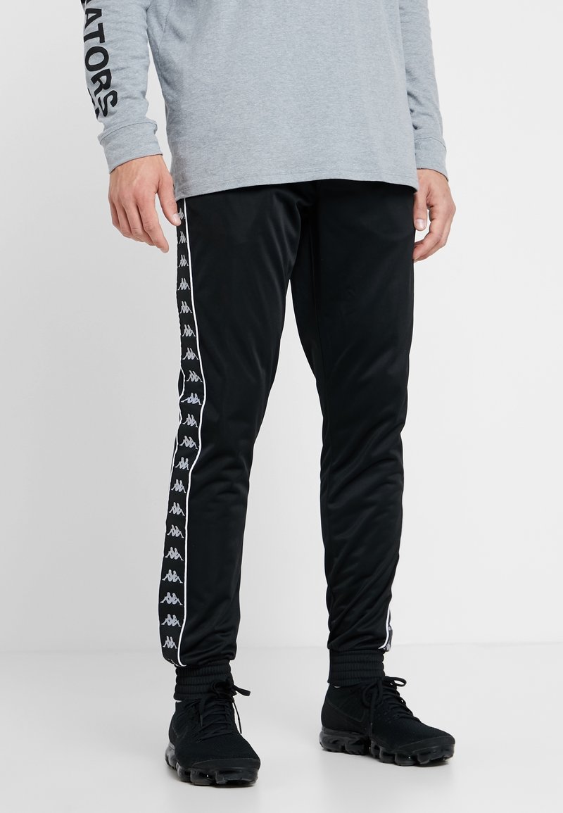 Svarta träningbyxor med elastiska muddar, med en vit sidremsa prydd med Kappa-logotyper. Tillverkade av ett mjukt, lätt tyg.