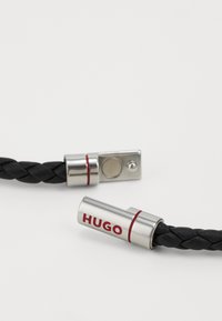 Svart flätad läderarmband med silverdetaljer som har "HUGO"-logotypen i röd färg. Spännet har ett cirkulärt knappdetalj.