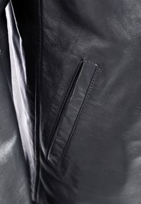 Schwarze Lederjacke mit glatter Textur, dezentem Glanz und einer rechteckigen Seitentasche. Das Innere ist gefüttert, was die Strapazierfähigkeit erhöht.