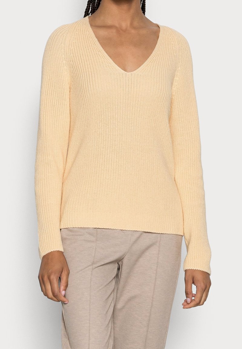 Femme portant un pull en maille beige clair col en V et un pantalon taupe clair, debout, les bras détendus le long du corps.