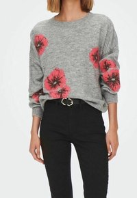 Grauer Strickpullover mit großen roten Blumenmustern, Rundhalsausschnitt und lockerer Passform. Kombiniert mit schwarzen Jeans und einer runden goldenen Gürtelschnalle.