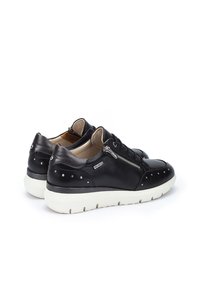 Zapatillas de cuero negro con un acabado suave, que presentan detalles de tachuelas metálicas, un zipper lateral y una suela de goma blanca.
