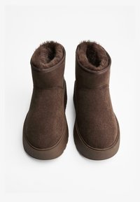 Un paio di stivaletti in suede marrone per l'inverno, con suole in gomma spesse e foderati in pelliccia morbida all'interno, mostrati dall'alto su uno sfondo bianco.