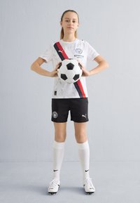 Baltas su raštais futbolo marškinėliai su juoda ir raudona įstrižaine juosta, juodi šortai, balti kojinės ir juodi futbolo bateliai. Laiko juodai baltą futbolo kamuolį.