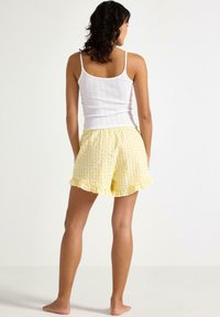 Hvid ribbet tanktop og gule gingham shorts med flæsekanter. Shortsene har en elastisk talje. Modellen ses fra ryggen.