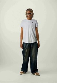 Vit bomullströja med en liten logotyp, kombinerad med löst sittande mörkblå denimjeans, bärs med ljusbruna slip-on skor, kort hår.