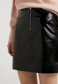 Mini-jupe en simili cuir noir avec une texture brillante, coutures visibles et fermeture éclair au dos. Associée à un pull rose.