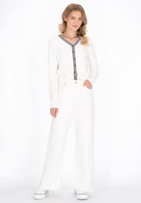 Witte gebreide top met een zwartgestreepte V-hals en knoopsluiting, gecombineerd met hoge, brede witte broeken en witte sneakers.