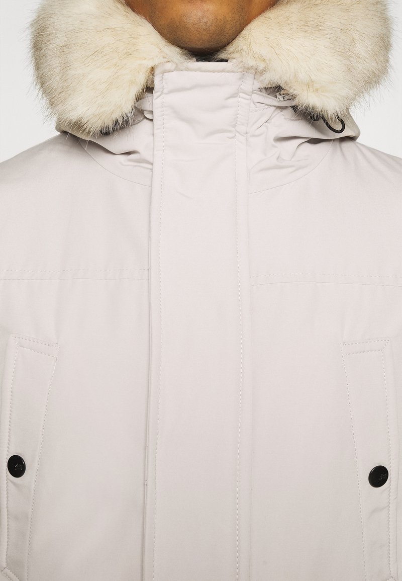 Veste beige clair avec un col en fausse fourrure, dotée d'une fermeture éclair et de boutons-pression, en tissu texturé et avec deux poches avant.