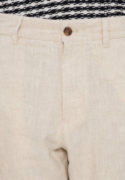 Nahaufnahme von beigen Leinenhosen mit einem einzelnen braunen Knopf und Gürtelschlaufen, getragen zu einem schwarz-weißen gemusterten Oberteil.