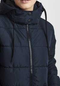Doudoune bleu marine avec col haut, fermeture éclair, capuche à cordon de serrage et texture matelassée prononcée. Dispose d'accents en métal noir.