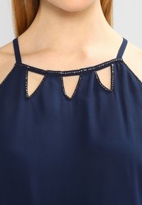 Kvinna som bär en marinblå ärmlös topp med pärlbesättning och utskurna droppformade former vid halsringningen.