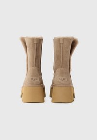 Bruine suède enkelboots met schapenwol voering, platte platformzool en logo-opdruk op de hak. De achterkant heeft een iets slouchy ontwerp.