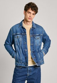 Chaqueta de mezclilla en azul medio con diseño clásico, cierres de botón y dos bolsillos frontales. Llevada sobre un suéter beige claro.