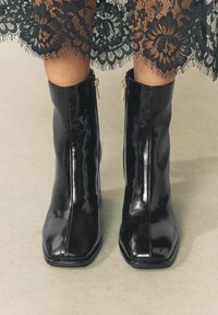 Czarne botki do kostki o błyszczącej, lakierowanej powierzchni, kwadratowym nosku i złotych suwaczkach po zewnętrznych stronach. Noszone z czarnym koronkowym dołem.