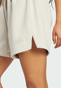 Světle béžové sportovní šortky se skluzem na boku, vyrobené z jemné tkaniny. Obsahují malé ražené logo a šité detaily.
