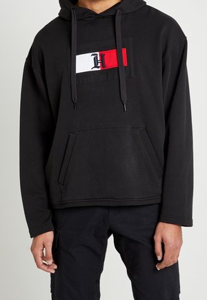 Mężczyzna w czarnej bluzie z kapturem z kieszenią z przodu oraz logo Tommy Hilfiger w kolorach czerwonym, białym i niebieskim na piersi, w połączeniu z czarnymi spodniami.