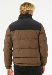 Bruine pufferjack met verticale stiksels, hoge kraag en een bovenste deel van donkerblauwe ribstof. De stof lijkt gestructureerd en licht van gewicht.