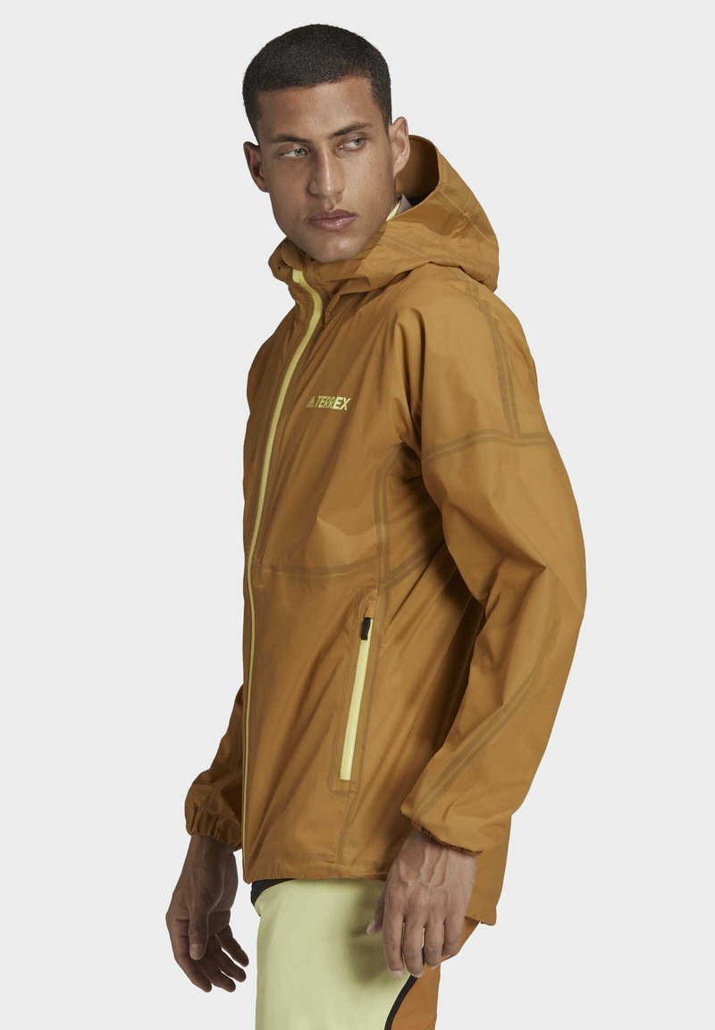 Chaqueta ligera marrón con capucha, con acentos y cremalleras en amarillo. Fabricada con un material suave y resistente al agua.