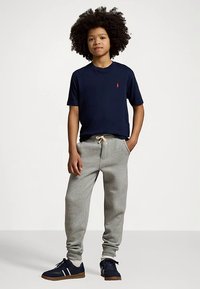 Młoda osoba z kręconymi włosami, ubrana w granatową koszulkę, szare dresy z troczkami i granatowe sneakersy w białe paski, stojąca z jedną ręką w kieszeni.