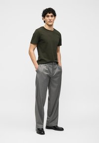 Jeune homme aux cheveux bouclés portant un t-shirt vert olive, un pantalon plissé gris, des chaussures noires, mains dans les poches, fond clair uni.