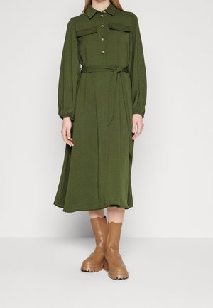 Vrouw draagt een olijfgroene midi-jurk met knopen, lange mouwen en een riem, gecombineerd met tankleurige grove halfhoge laarzen, staand tegen een witte achtergrond.