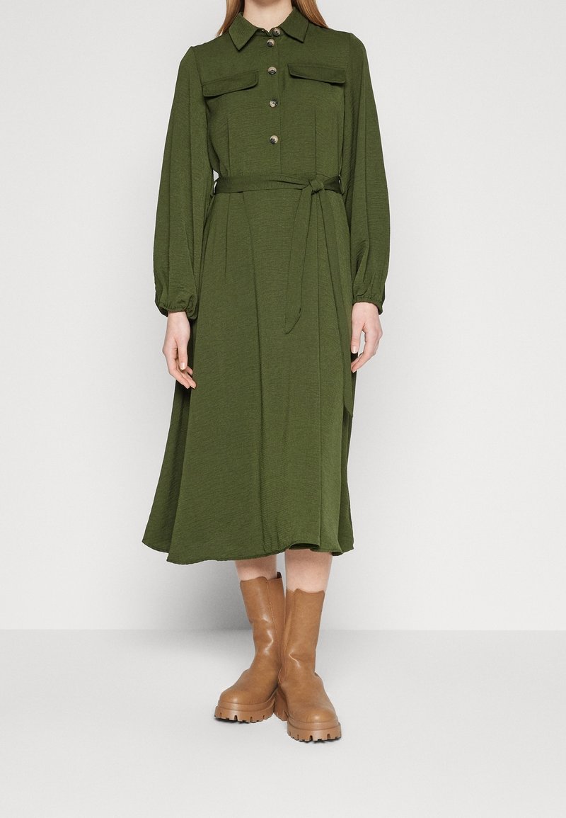 Femme portant une robe midi boutonnée vert olive à manches longues et ceinture, associée à des bottes épaisses couleur tan arrivant mi-mollet, debout sur fond blanc.