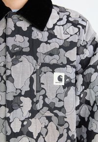 Giacca camouflage nera e grigia con colletto in velluto nero, bottoni a pressione e logo Carhartt sulla tasca sinistra del petto.