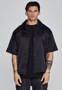 Camisa negra de manga corta con patrón texturizado, botonadura frontal y un cuello amplio, superpuesta a una camiseta negra. Ajuste casual, tejido ligero.