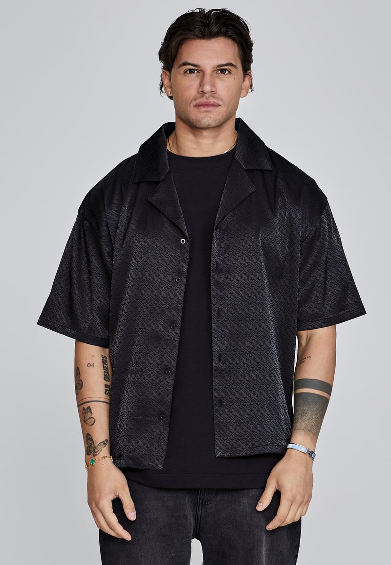 Camisa negra de manga corta con patrón texturizado, botonadura frontal y un cuello amplio, superpuesta a una camiseta negra. Ajuste casual, tejido ligero.
