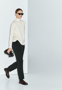 Donna che cammina indossando un maglione a collo alto color crema, pantaloni neri, mocassini scuri, occhiali da sole e tiene in mano una borsa a tracolla nera intrecciata.