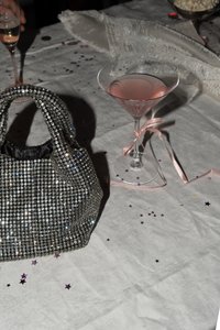 Czarna i srebrna torebka zdobiona koralikami, o teksturowanej powierzchni i zaokrąglonych uchwytach, obok różowego koktajlu w przezroczystej szklance, ustawiona na stole.