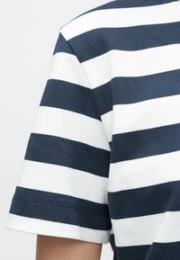 Tamsiai mėlynas ir baltas dryžuotas marškinėliai su apykakle ir trumpomis rankovėmis. Audinys glotnus, matomas mezgimo raštas.